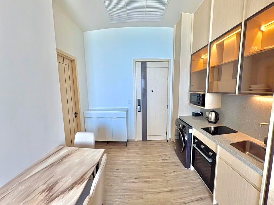 Modern kitchen with light wood cabinets, stainless sink, oven, stove, and a small dining table with two chairs along the left wall.