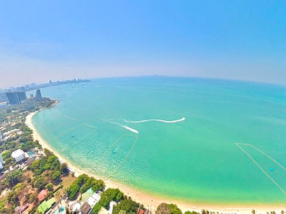 Aerial view of a curved sandy beach along a turquoise bay with a city skyline on the left and green palm trees in the foreground.