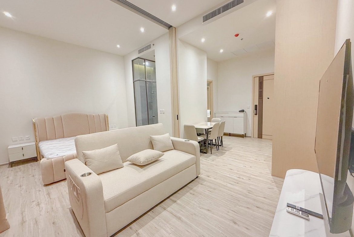Bright, modern studio living area with a beige sofa, cushions, a bed in the back, and a dining table with six chairs in a neutral-toned space.