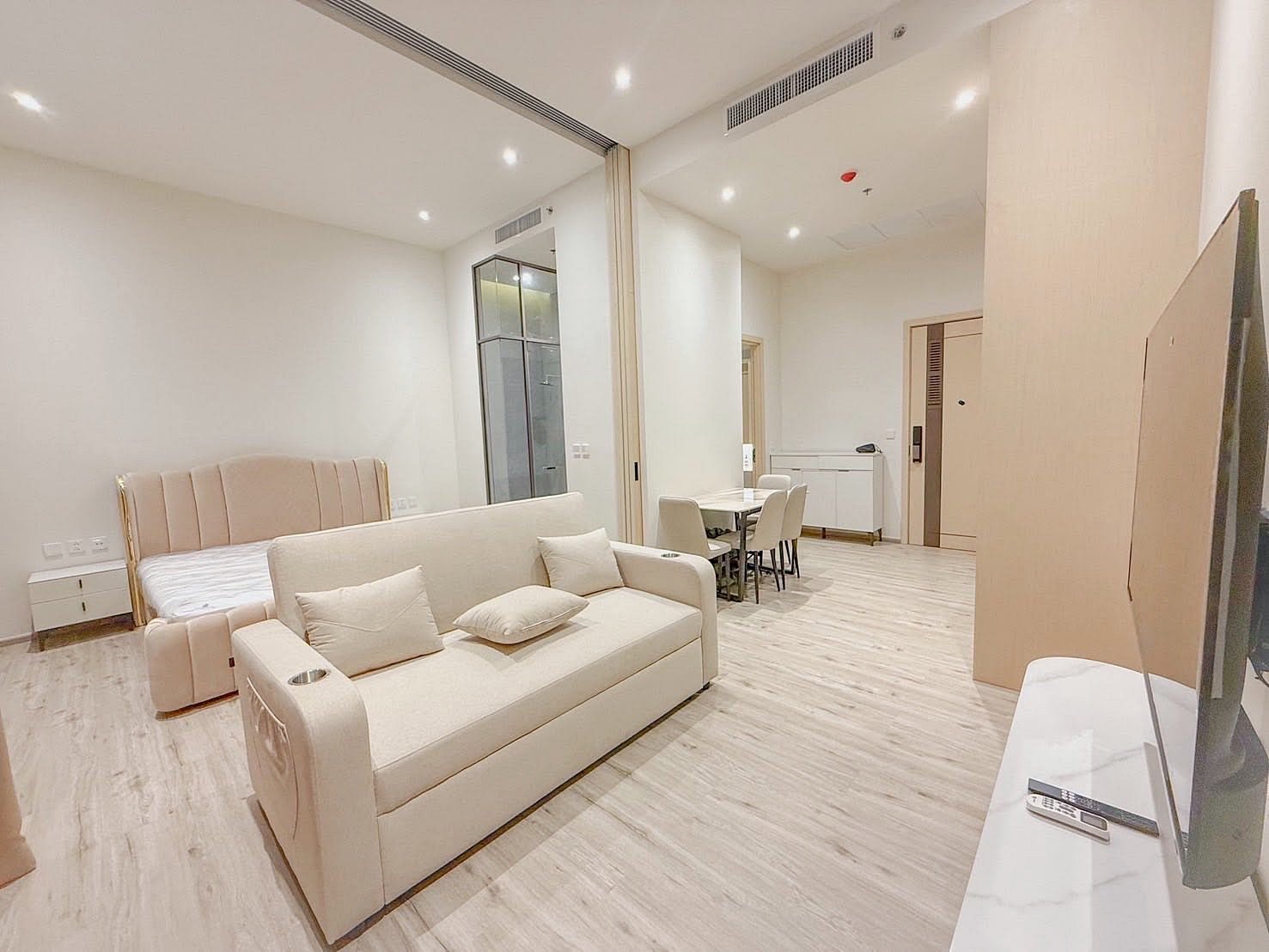 Arom_Wongamat_5005_6 Bright, modern studio living area with a beige sofa, cushions, a bed in the back, and a dining table with six chairs in a neutral-toned space.