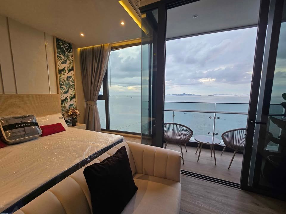 Hotel room with a large bed, beige headboard, and a sofa, sliding glass doors open to a balcony overlooking the calm sea and distant islands on a cloudy day.