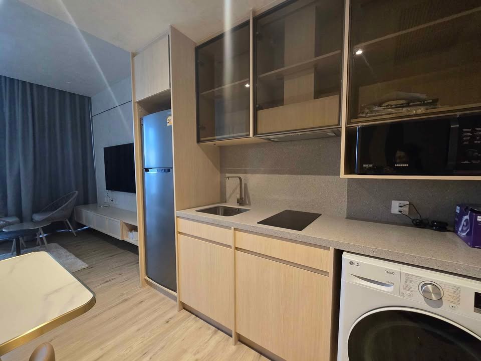 Compact kitchen with light wood cabinets, stainless fridge, sink, induction cooktop, microwave, and washing machine visible nearby.