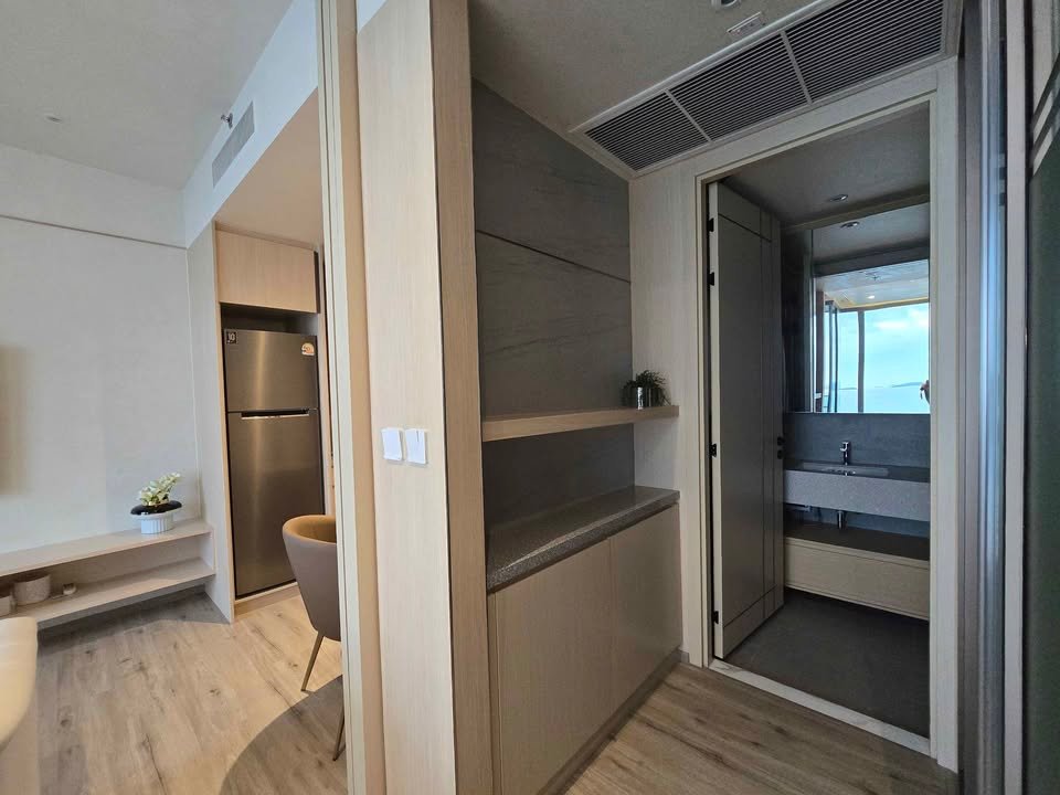 Open-plan living area with a kitchenette, stainless fridge, and built-in shelves leading to a hallway and a bathroom visible in the distance.