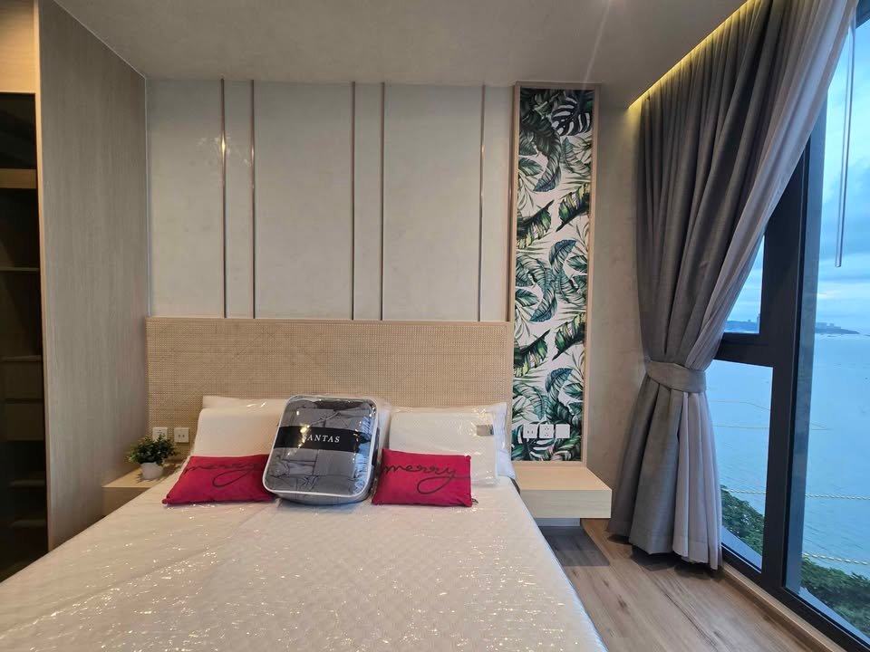 Modern hotel bedroom with a king bed, beige upholstered headboard, twin nightstands, and tropical leaf wallpaper panel on the wall beside the bed; gray curtains frame a large window with an ocean view; a suitcase and two pink pillows sit on the bed with a small plant on the left nightstand.
