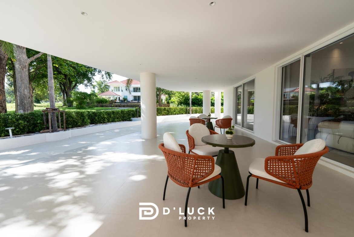 Covered outdoor seating area with orange wicker chairs around round green tables, next to glass sliding doors and a landscaped garden.
