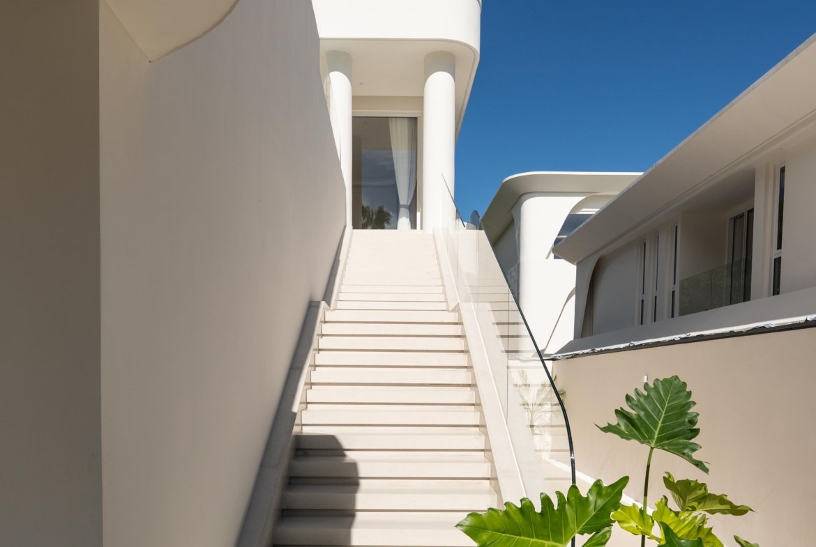 White modern building exterior with a central flight of stairs, curved balconies, and glass railings, set against a clear blue sky; tropical leaves frame the foreground.