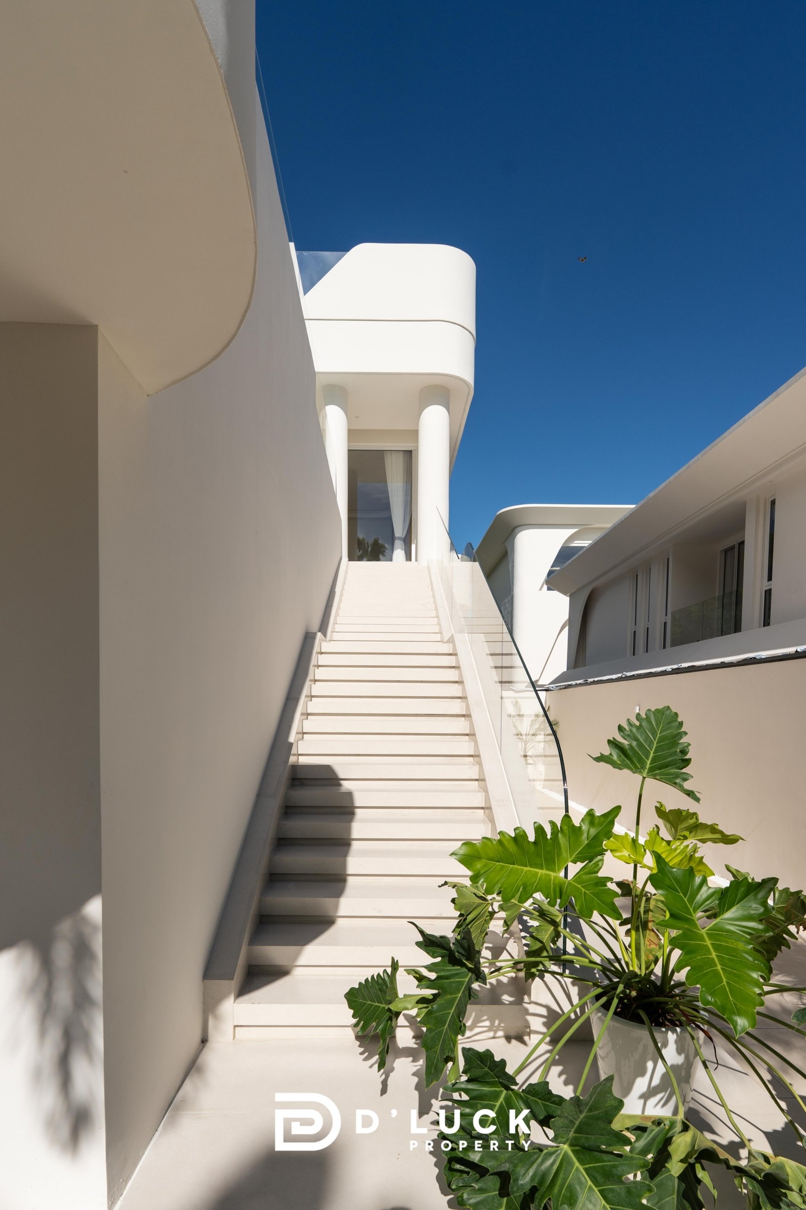 Big_V_Pattaya_Type_A_127 White modern building exterior with a central flight of stairs, curved balconies, and glass railings, set against a clear blue sky; tropical leaves frame the foreground.