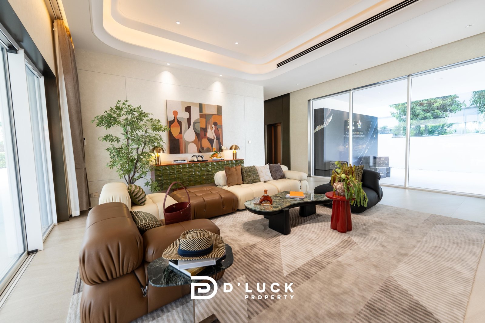 Big_V_Pattaya_Type_A_130 Contemporary living room with a cream leather sectional, abstract art on the wall, a tall green plant, and floor-to-ceiling windows letting in light.