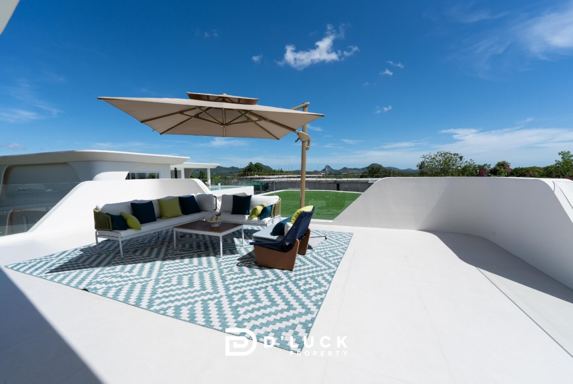 Rooftop lounge with white outdoor sofa, teal and lime cushions, a large beige umbrella, and a patterned teal‑and‑white rug under bright blue skies.