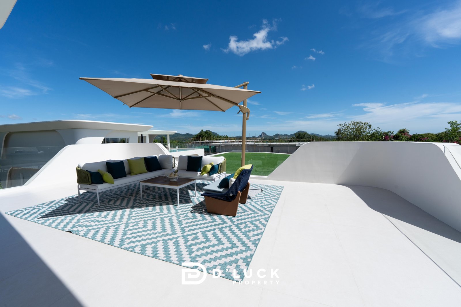 Big_V_Pattaya_Type_A_16 Rooftop lounge with white outdoor sofa, teal and lime cushions, a large beige umbrella, and a patterned teal‑and‑white rug under bright blue skies.
