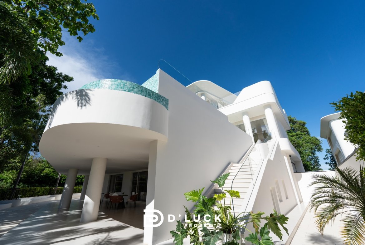 Modern white villa with curved balconies and turquoise tile accent, set against a clear blue sky with surrounding tropical greenery.