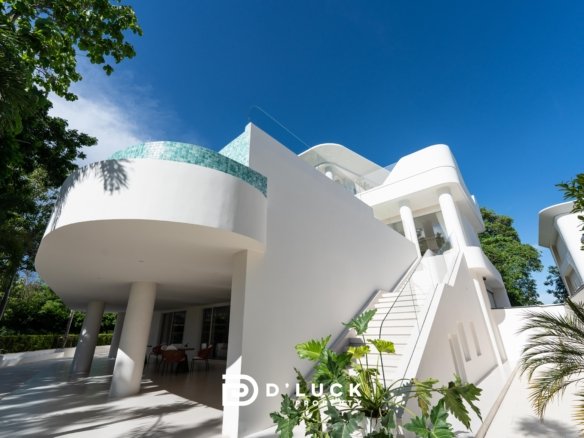 Modern white villa with curved balconies and turquoise tile accent set against a clear blue sky with surrounding tropical greenery