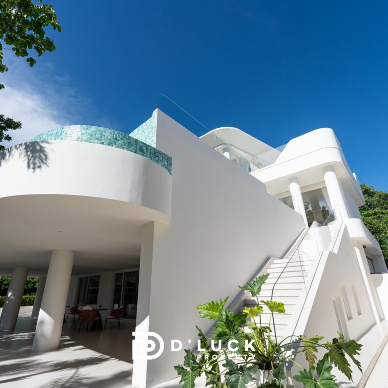 Modern white villa with curved balconies and turquoise tile accent set against a clear blue sky with surrounding tropical greenery