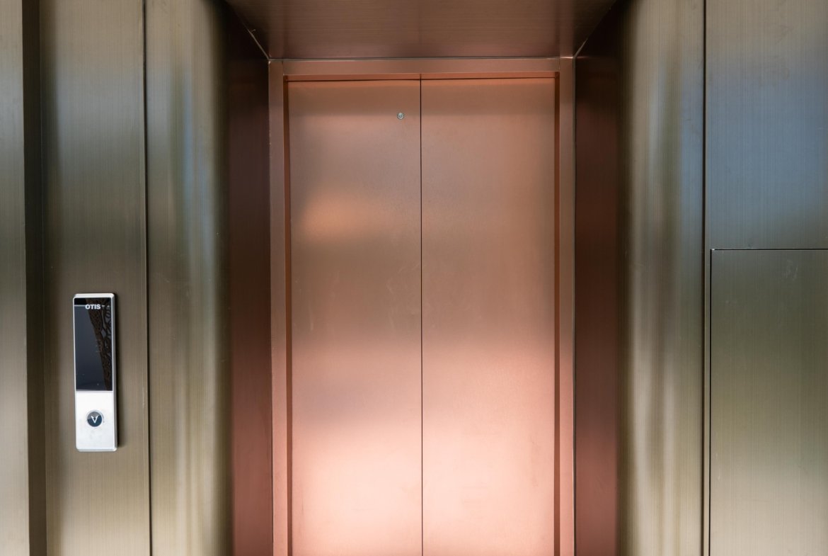 Copper elevator doors set in dark metal walls, with a left-side control panel and the D'LUCK Property logo on the floor in front of the doors.