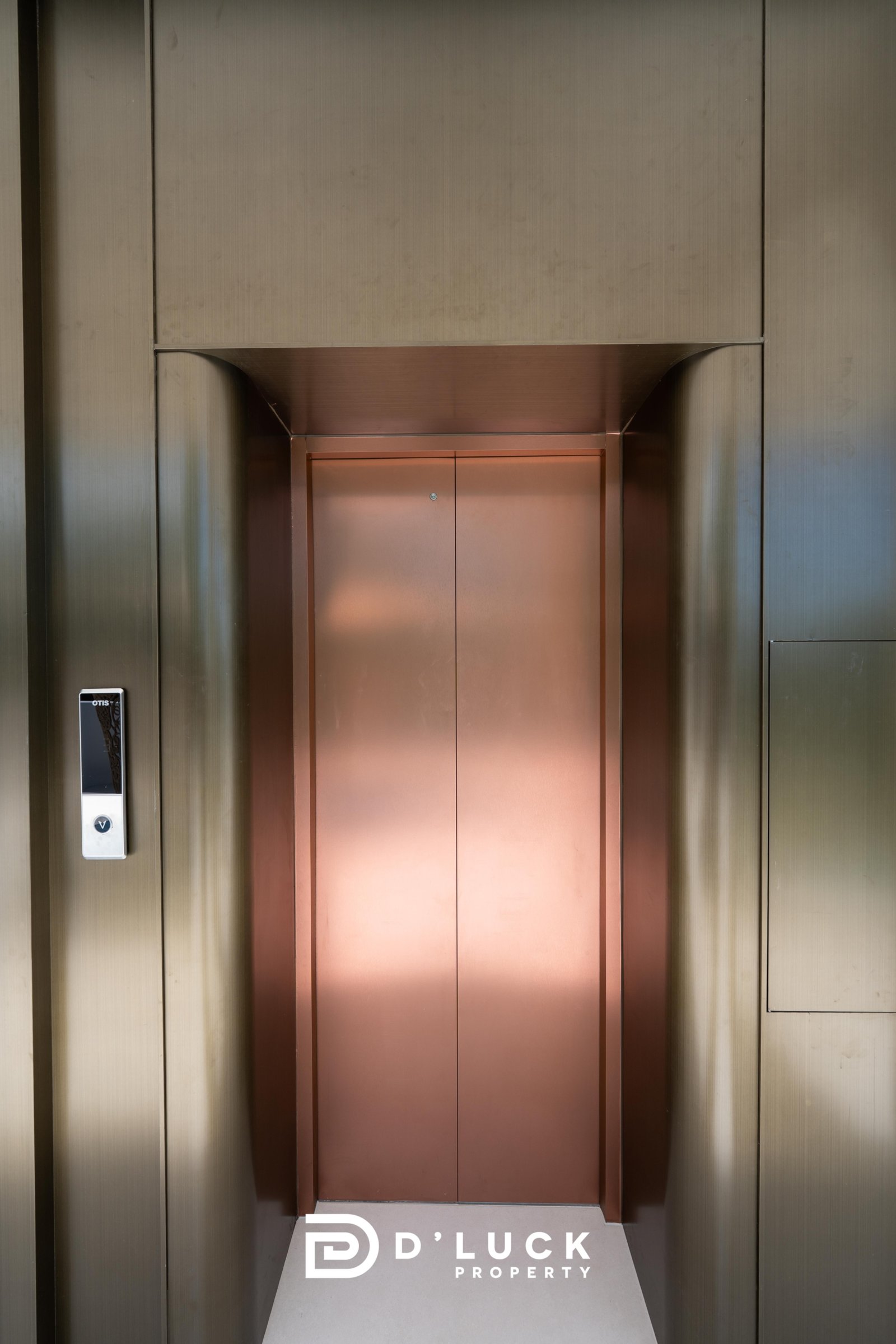 Big_V_Pattaya_Type_A_23 Copper elevator doors set in dark metal walls, with a left-side control panel and the D'LUCK Property logo on the floor in front of the doors.