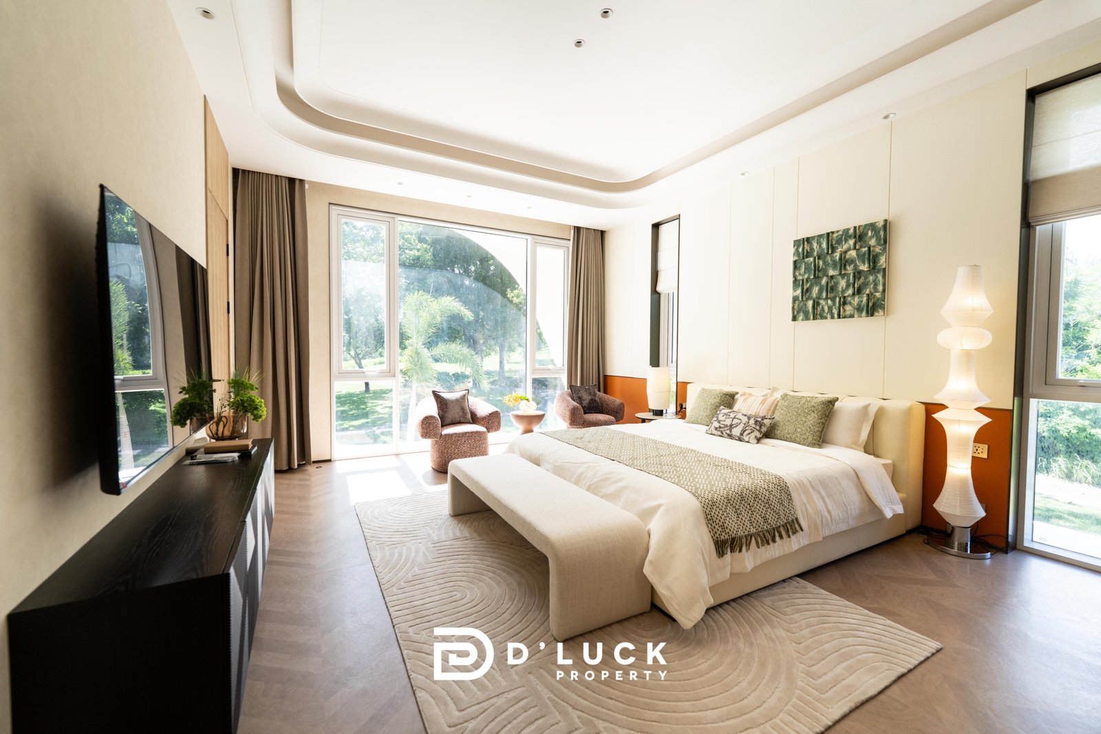 Big_V_Pattaya_Type_A_45 Luxurious hotel bedroom with a king bed, beige tones, curved white ceiling, and a seating area by large windows overlooking greenery; D'LUCK PROPERTY logo at foot of bed.