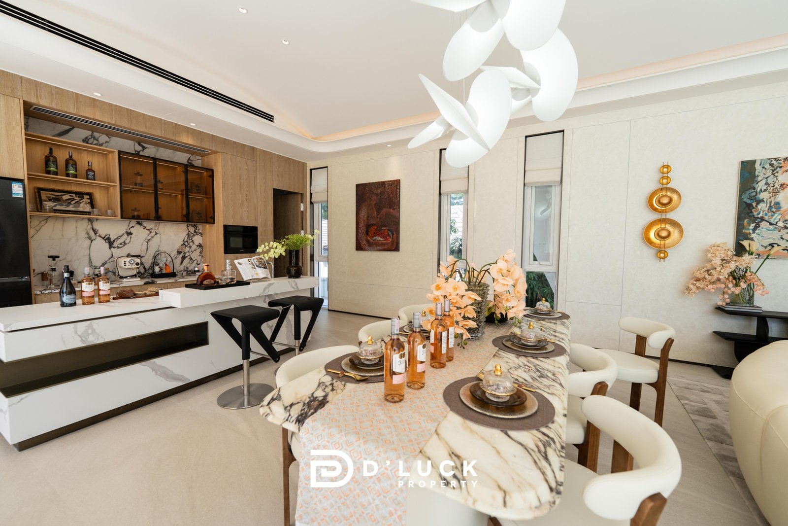 Big_V_Pattaya_Type_A_5 Open-plan kitchen and dining space with marble surfaces, wooden cabinets, and a sculptural white chandelier; bottles and food on the island hint at a party setup.