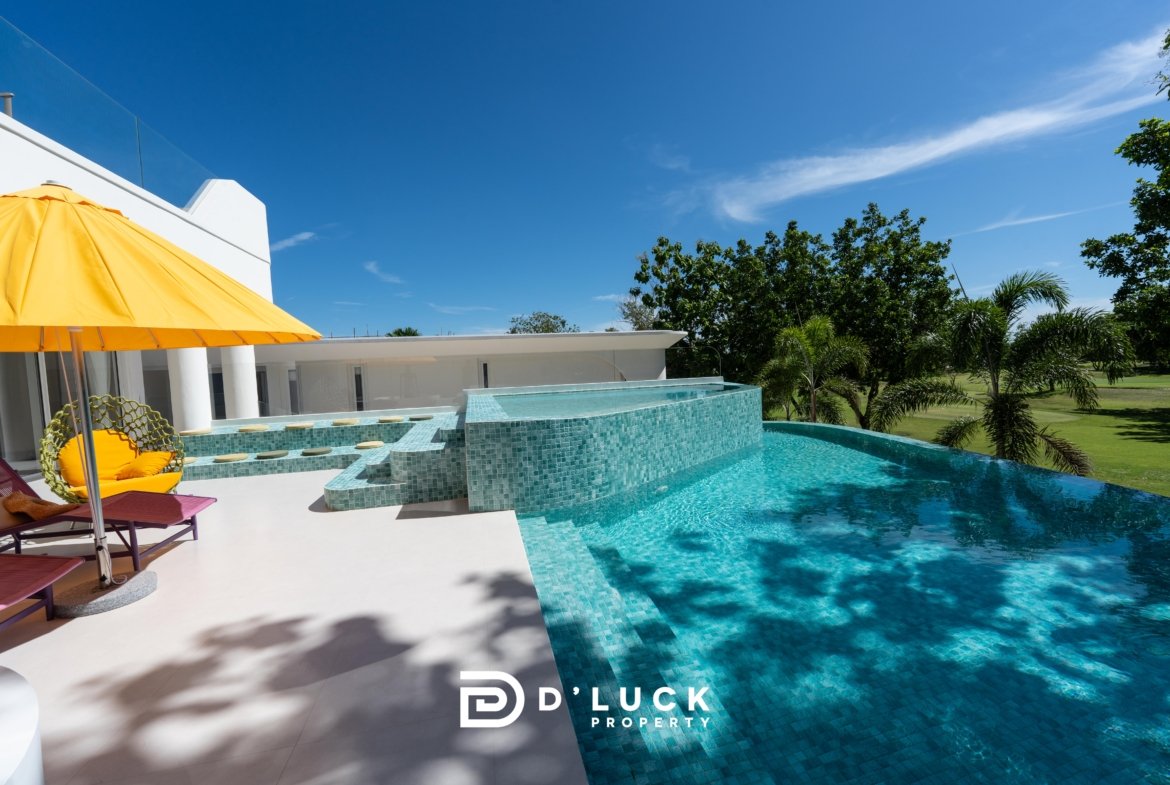 Modern white villa with a bright yellow patio umbrella, turquoise mosaic pool, and sun loungers by the water under a clear blue sky.