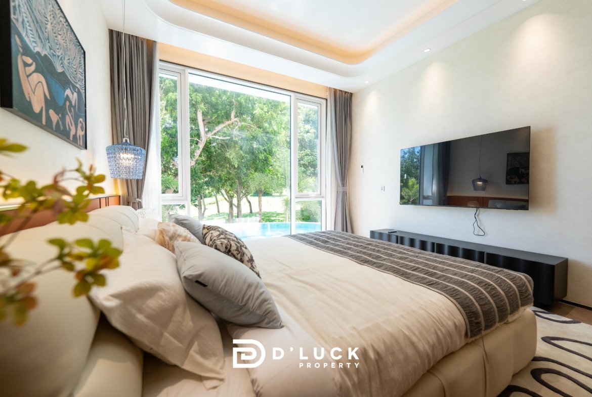 Modern bedroom with a large beige bed, gray striped bedding, a wall-mounted TV, and tall windows overlooking a lush garden.