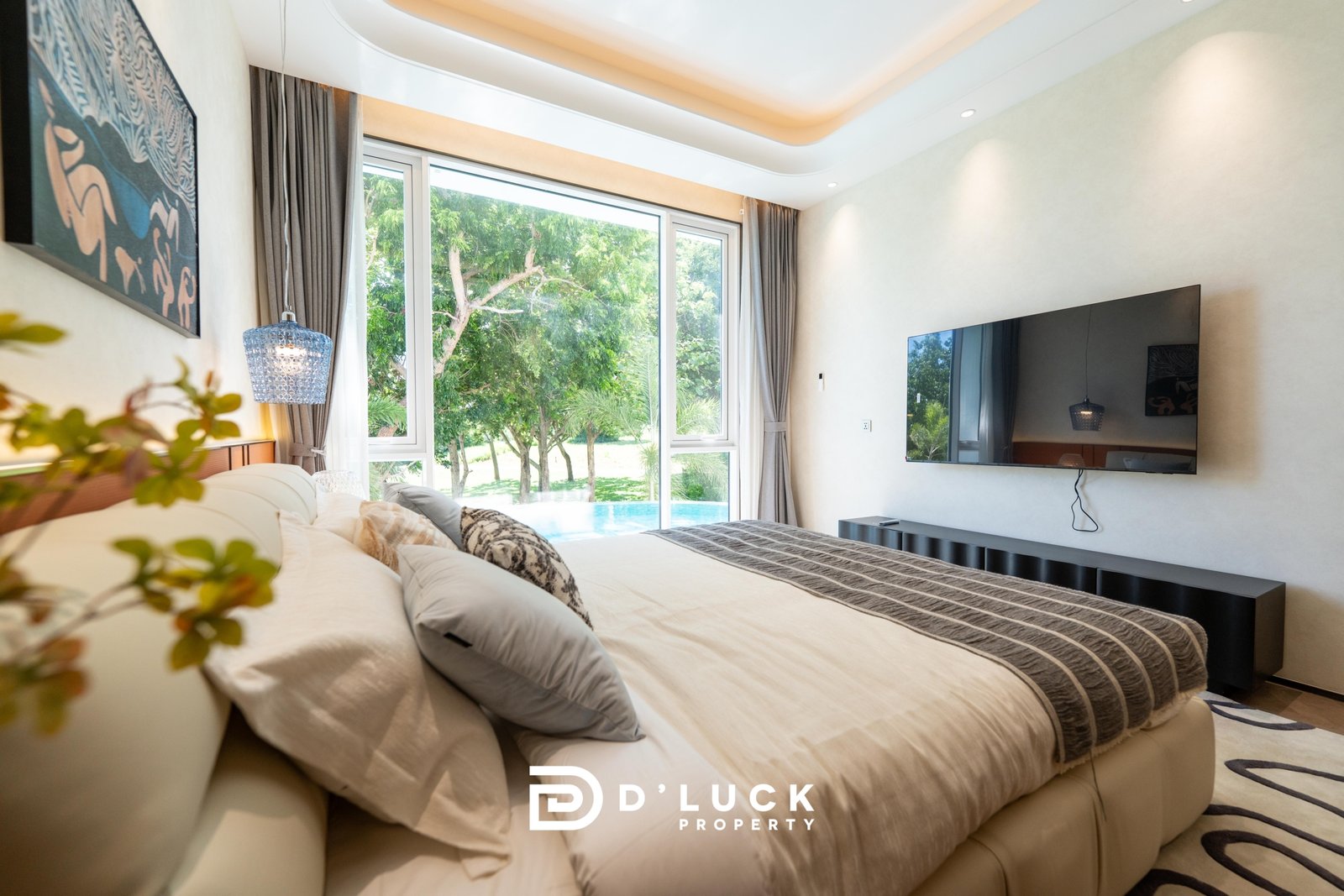Big_V_Pattaya_Type_A_81 Modern bedroom with a large beige bed, gray striped bedding, a wall-mounted TV, and tall windows overlooking a lush garden.