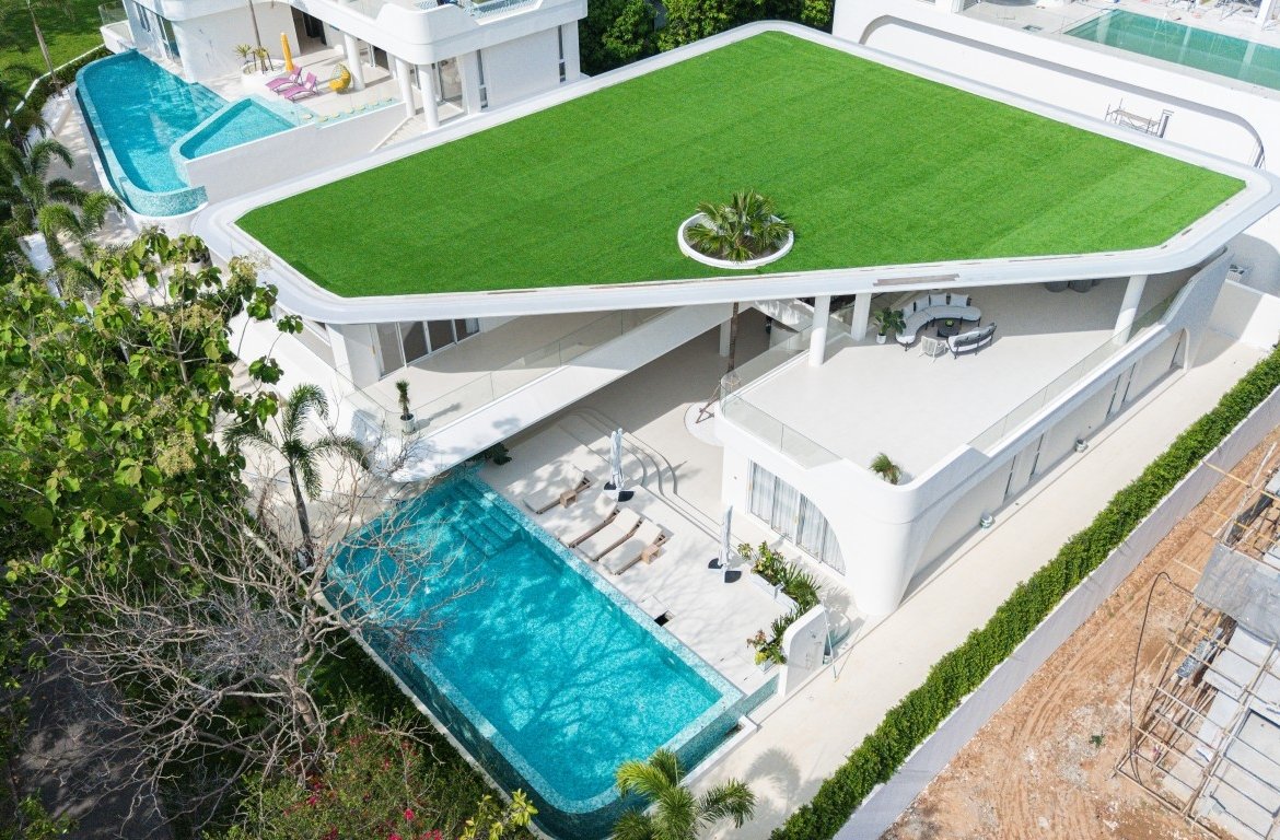 Aerial view of a modern white villa with a green rooftop lawn, circular planter, and turquoise pool nearby.