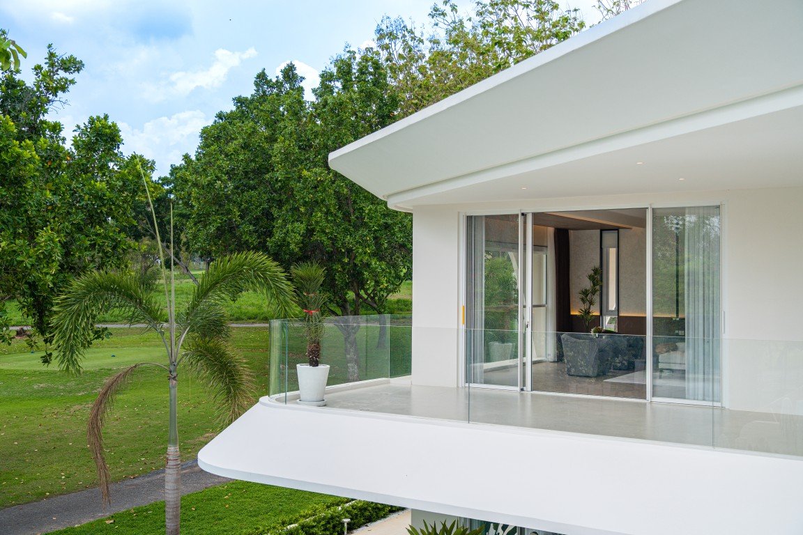 Modern white house balcony with glass railing, open sliding doors revealing a living room with seating, overlooking a green lawn and palm trees on a sunny day.
