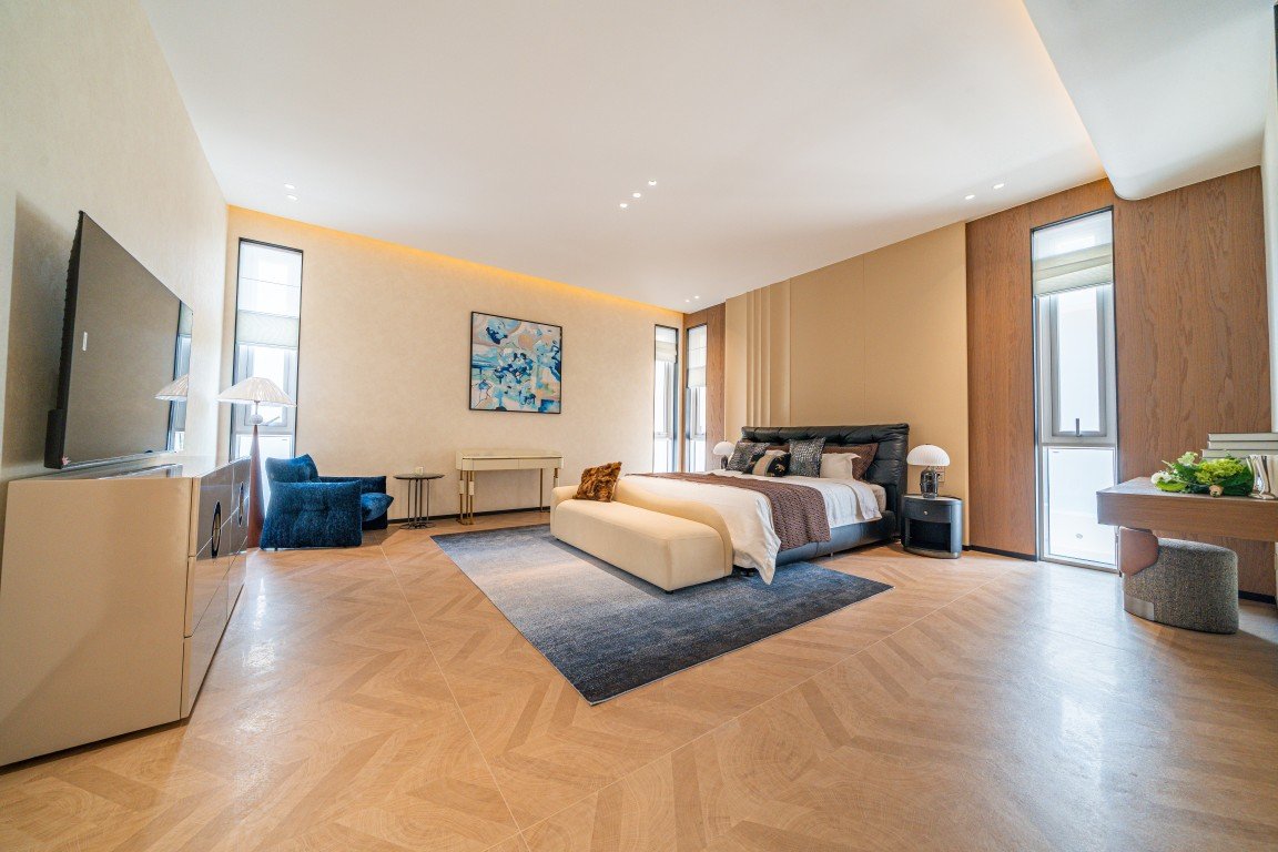 Spacious luxury bedroom with a king bed, beige bench at the foot, dark headboard, and a blue area rug under the sleeping area.