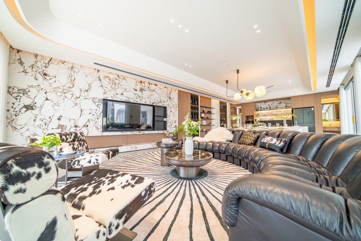 Spacious modern living room with a black leather sectional, cowhide chairs, circular rug, and a marble feature wall behind a flat-screen TV.
