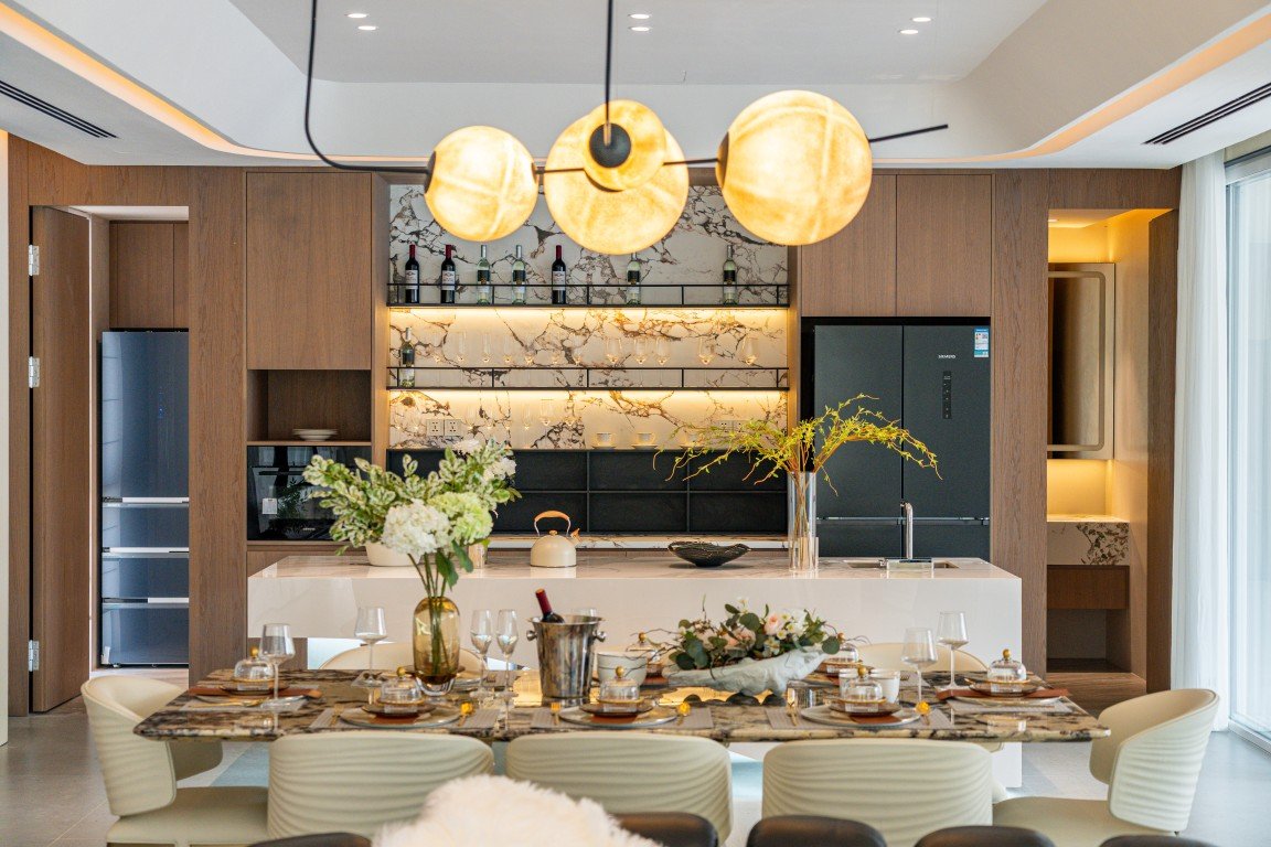 Modern open-plan kitchen and dining area with marble backsplash, a long wooden table set for a formal meal, and warm pendant lights overhead, ready for entertaining.