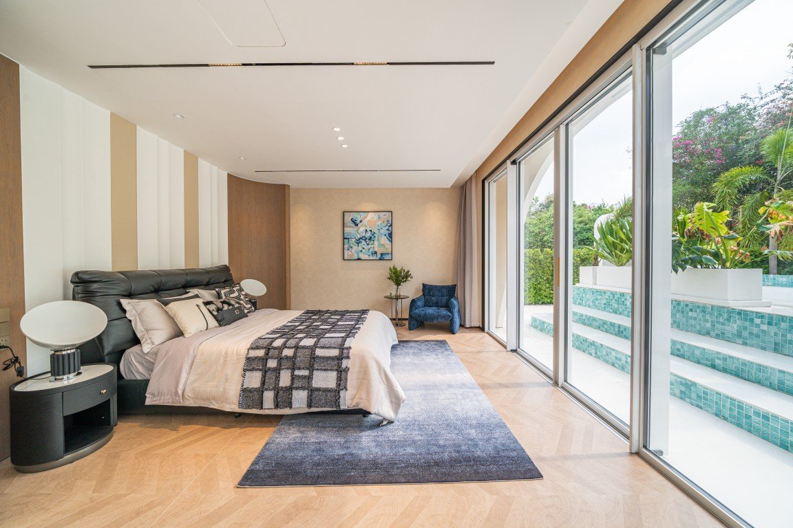 Spacious modern bedroom with a king bed, dark upholstered headboard, nightstands, and a blue rug beside floor‑to‑ceiling glass doors. Outdoors shows a pool and greenery.