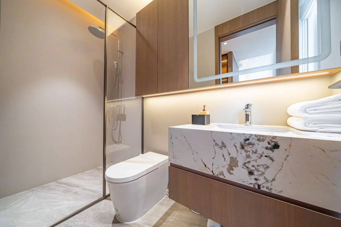 Modern bathroom with glass-enclosed shower, wall-mounted rainfall showerhead, and marble vanity top with stacked towels and chrome faucet.