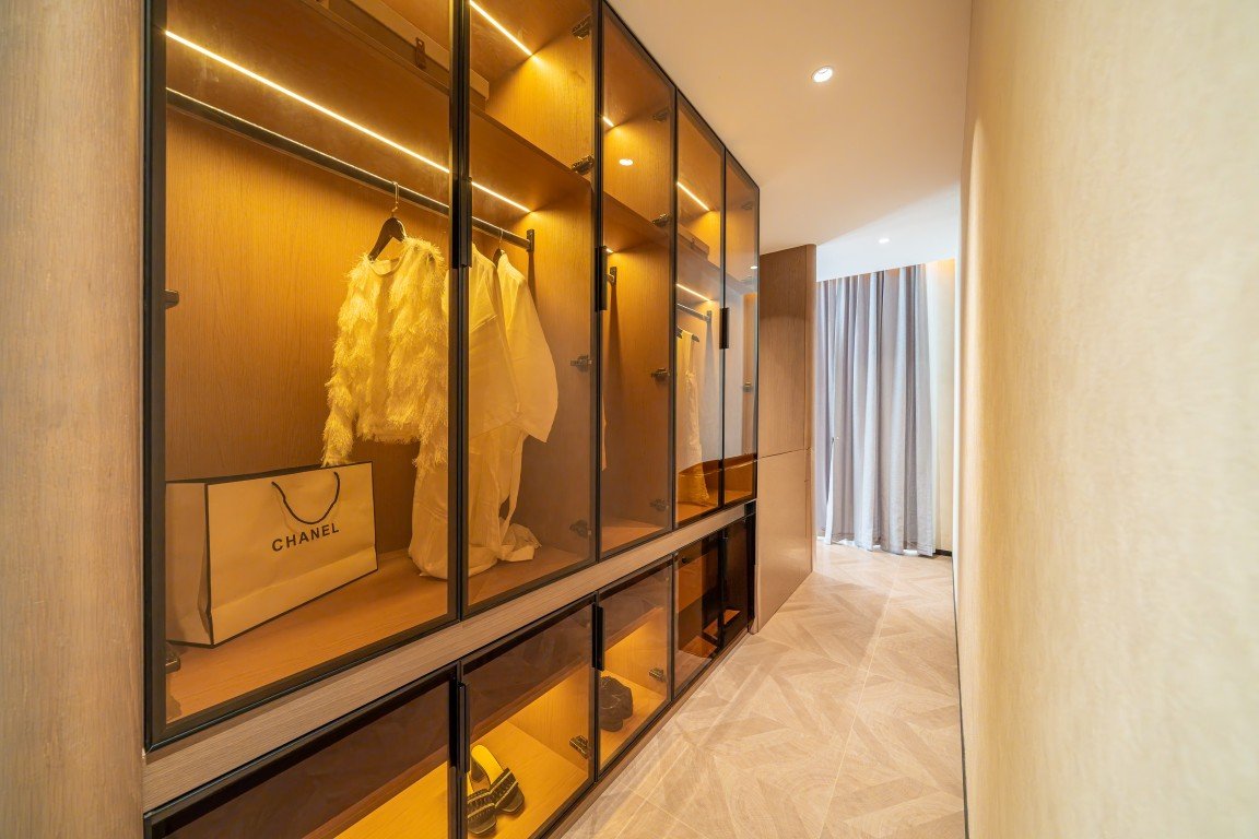 Walk-in closet with glass-front wardrobes, warm lighting, dresses hanging, and a Chanel shopping bag on a shelf inside.