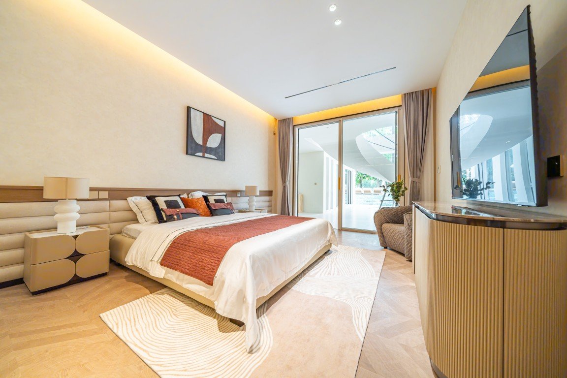 Hotel bedroom with a king bed, beige headboard, orange quilt and plush cushions, flanked by nightstands and lamps; sliding glass door to a balcony in the background.