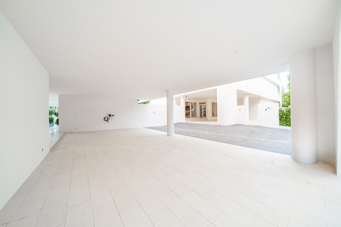 Bright, minimalist white courtyard with large tiled floor, a single column, and an entryway to a modern building in the background, daytime light.