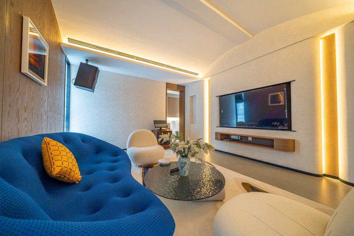 Modern living room with a bright blue tufted sofa, yellow pillow, and round glass coffee table facing a wall-mounted flat-screen TV. Warm ambient lighting and white seating create a contemporary vibe.