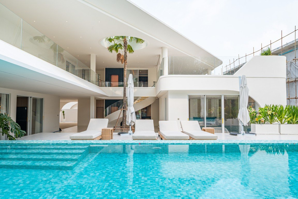 Luxurious white hotel villa with a turquoise swimming pool and row of white lounge chairs on a pool deck under a clear sky