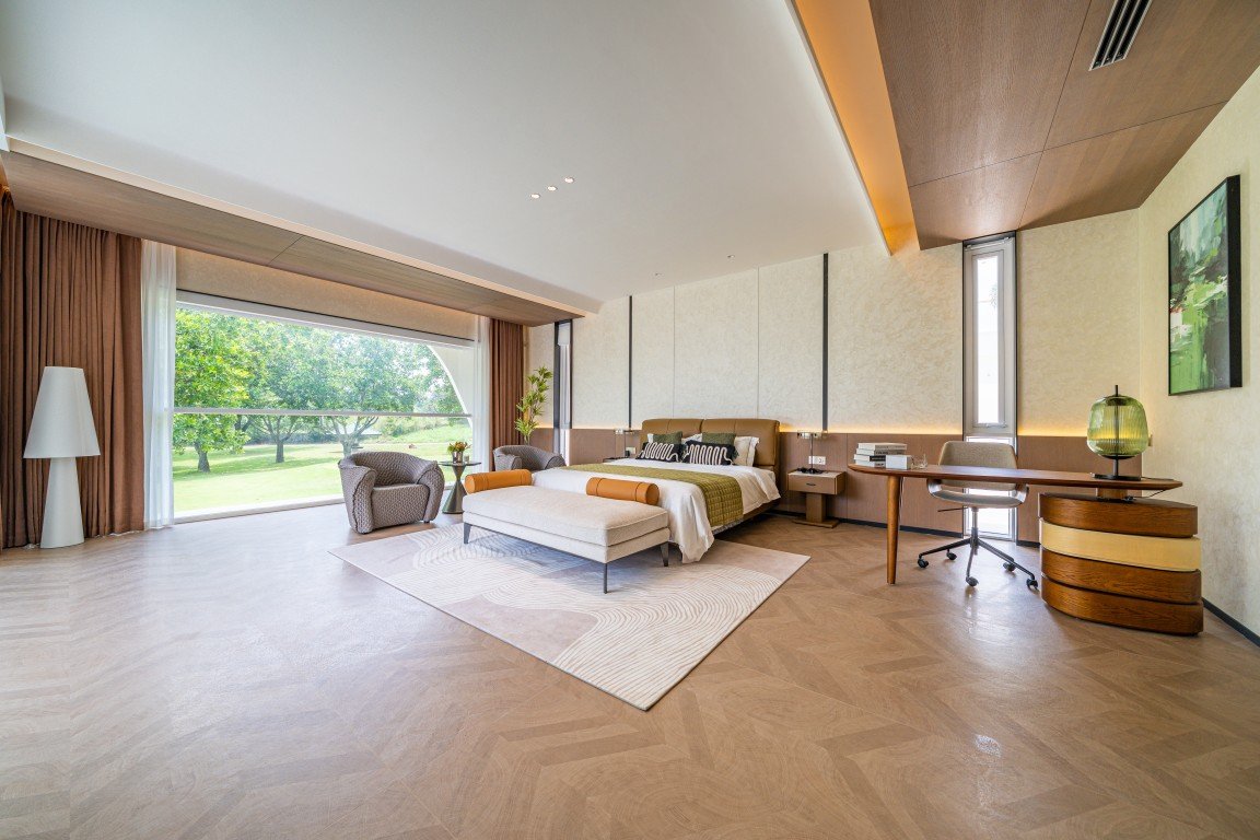 Luxurious hotel suite with a king bed, ottoman, and lounge chairs, plus a desk and a large window overlooking greenery.