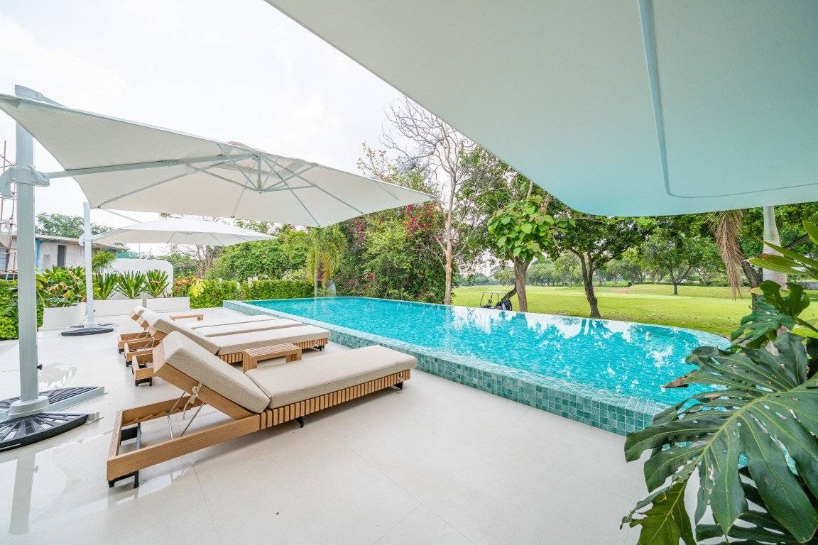 Outdoor poolside with beige loungers and large white umbrellas on a light deck, lush greenery in the background.