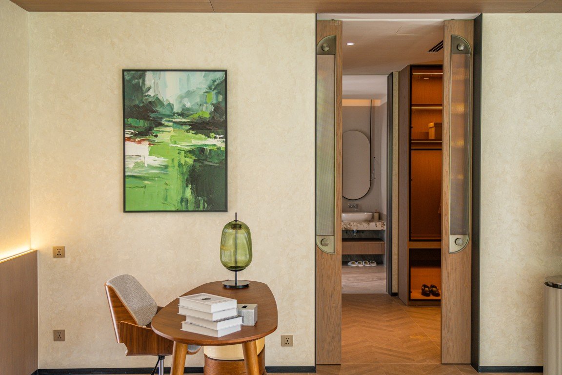 A round wooden desk with a stack of white books, a green glass lamp, and a cushioned chair in a softly lit hotel room, with abstract green artwork on the wall behind.