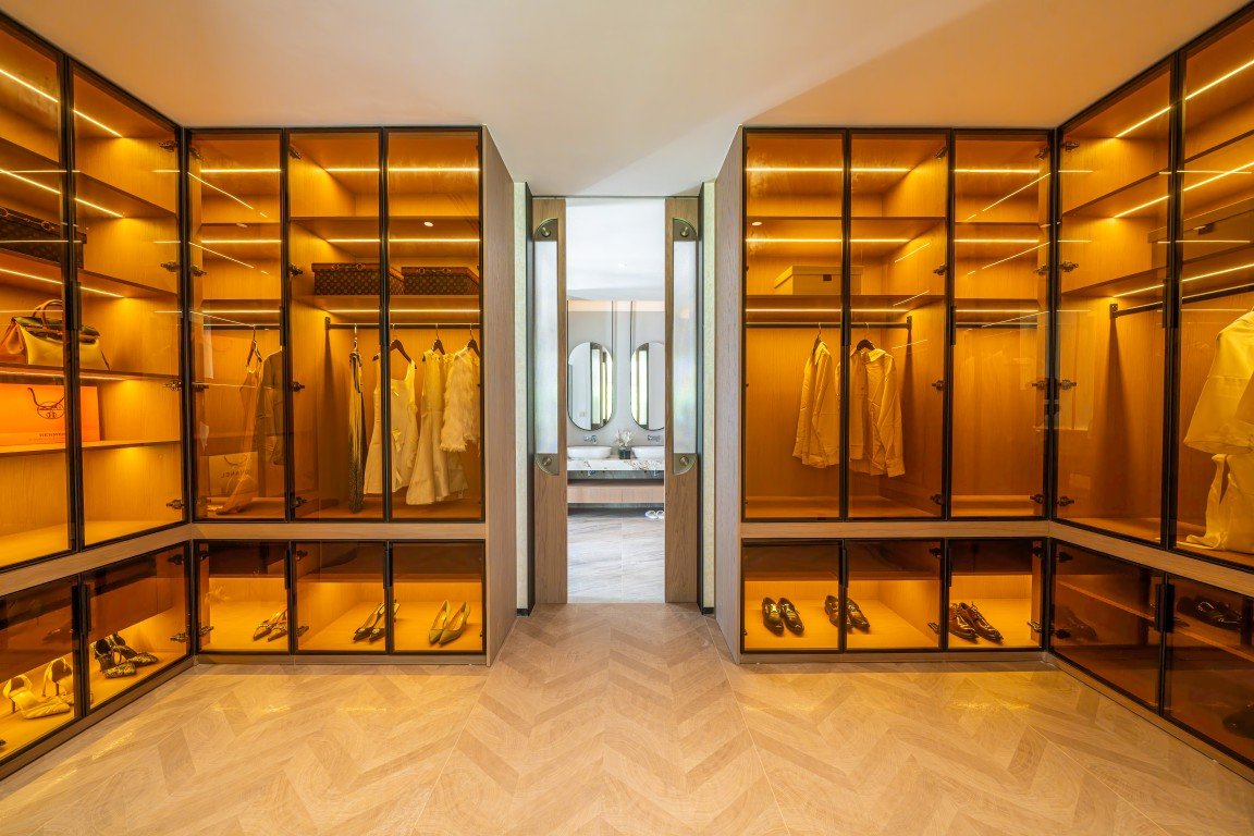 Walk-in luxury closet with glass-front wardrobes, warm amber lighting, and shoes displayed on shelves.