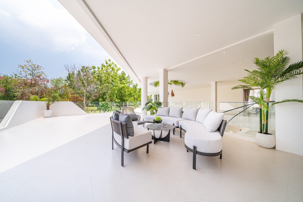 Modern outdoor lounge on a bright terrace with a curved white sectional sofa and potted plants nearby.