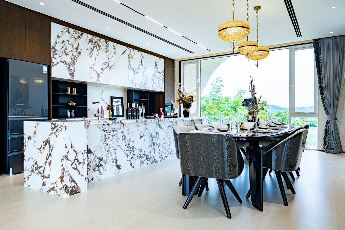 Luxurious open kitchen with a marble island and built-in storage, next to a round dining table set for six by a large window.
