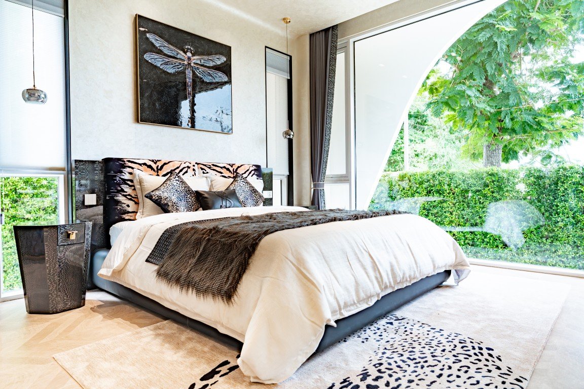 Luxurious modern bedroom with a low bed, white duvet, animal-print pillows, and a large glass wall opening to greenery outside