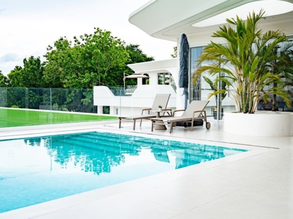 Modern white villa with a rectangular blue pool and lounge chairs on a sunlit terrace glass railing and tropical plants nearby