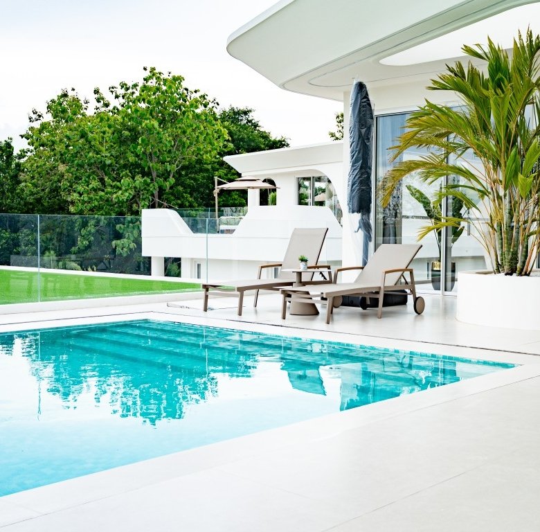 Modern white villa with a rectangular blue pool and lounge chairs on a sunlit terrace glass railing and tropical plants nearby