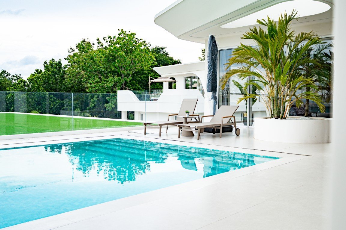 Modern white villa with a rectangular blue pool and lounge chairs on a sunlit terrace glass railing and tropical plants nearby