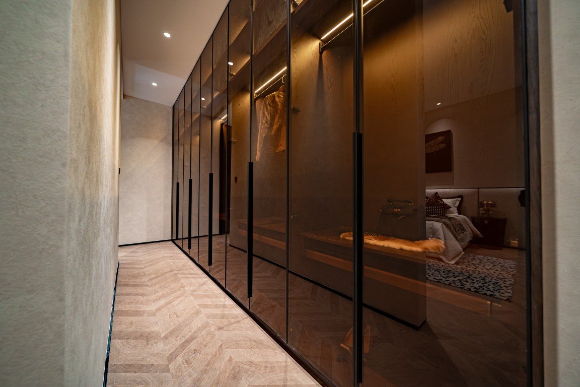 Long glass-walled wardrobe corridor with warm lighting, revealing a bedroom through the panes.