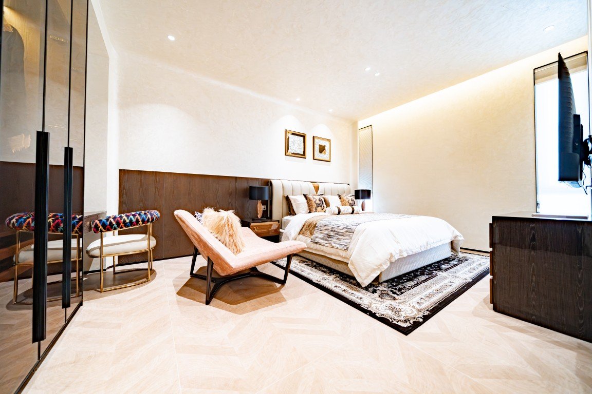 Luxurious hotel bedroom with a king bed, upholstered headboard, and a pink lounge chair by the foot of the bed, a black-and-white rug, and wall-mounted lamps.