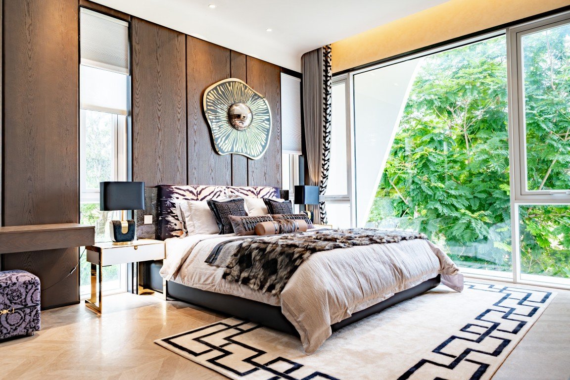 Luxurious modern bedroom with dark wood paneled wall, king-size bed, and floor-to-ceiling windows overlooking greenery, plus a fur throw and decorative pillows.