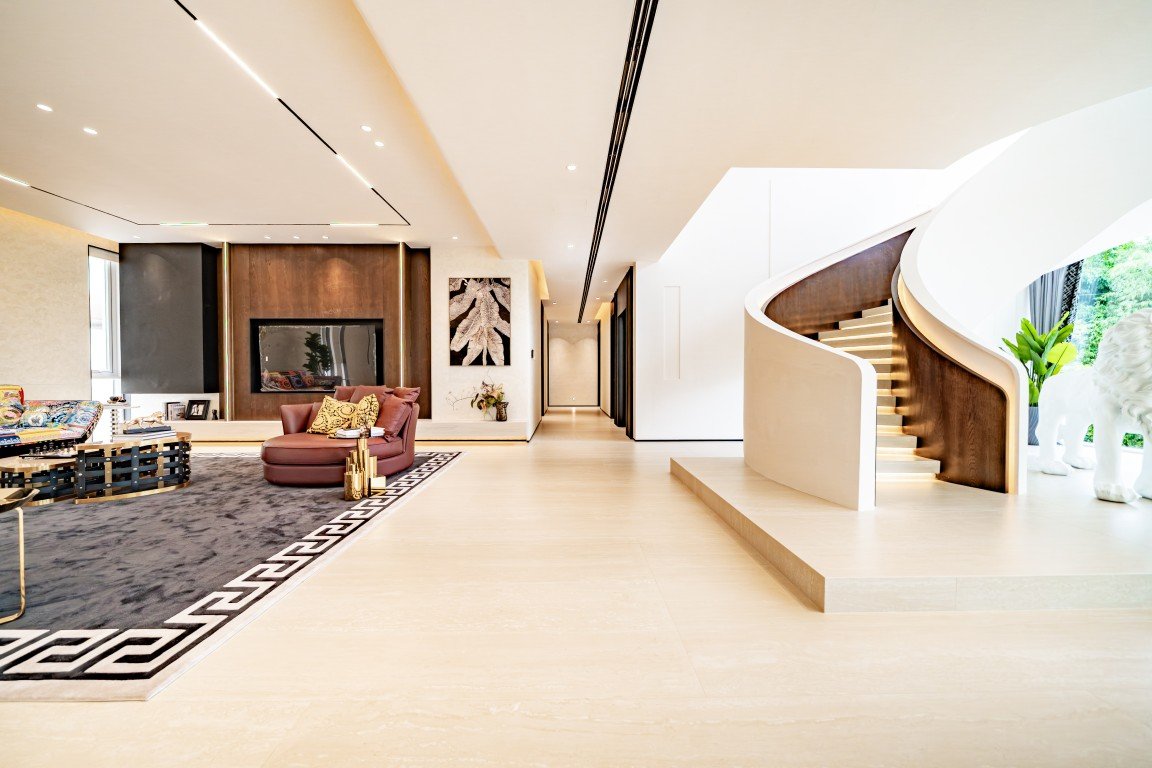 Luxurious open living area with a curved white spiral staircase, cream floors, and a burgundy sofa by a dark rug.